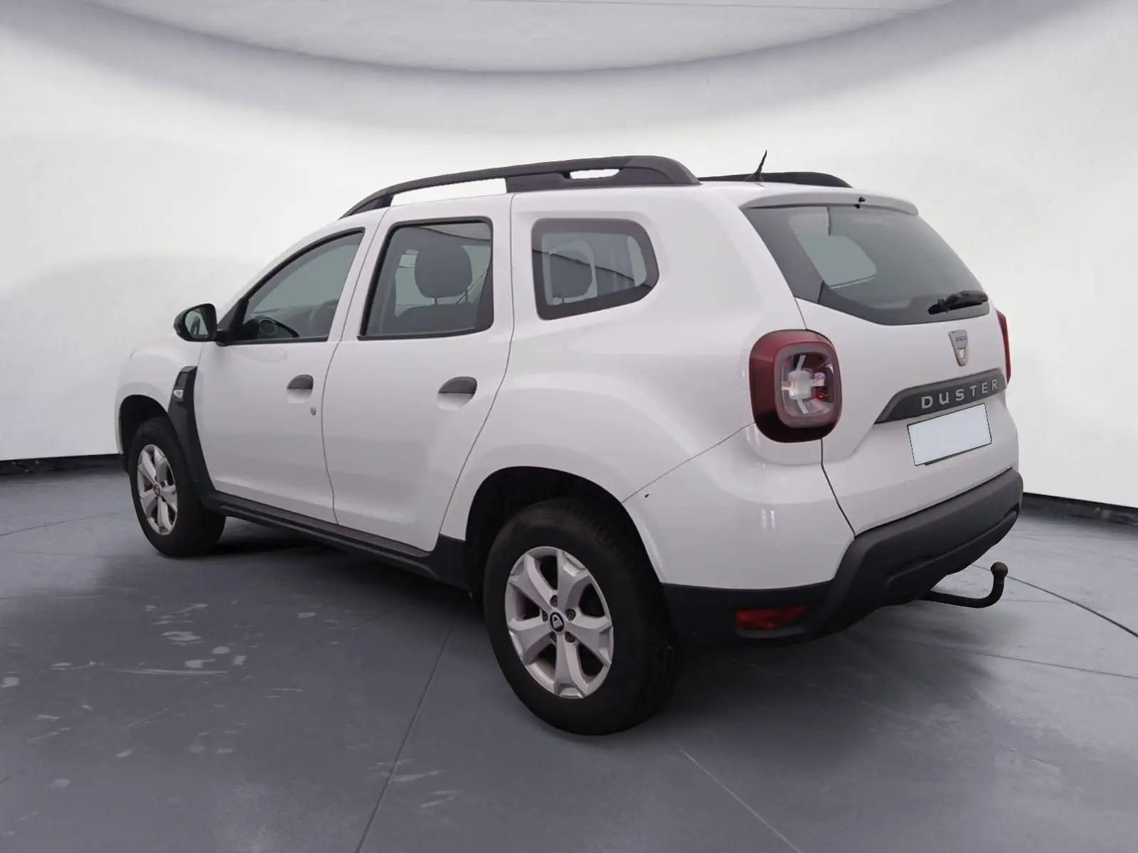 Vue 3/4 arrière droite d’un Dacia Duster blanc 2021 avec attelage et barres de toit noires visibles.