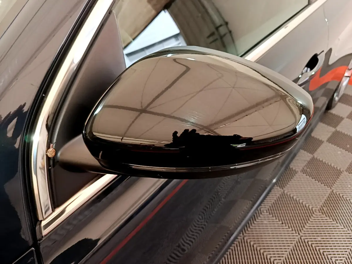 Gros plan sur le rétroviseur côté gauche noir brillant d'une Peugeot 308 gris foncé, reflet de la structure environnante visible.
