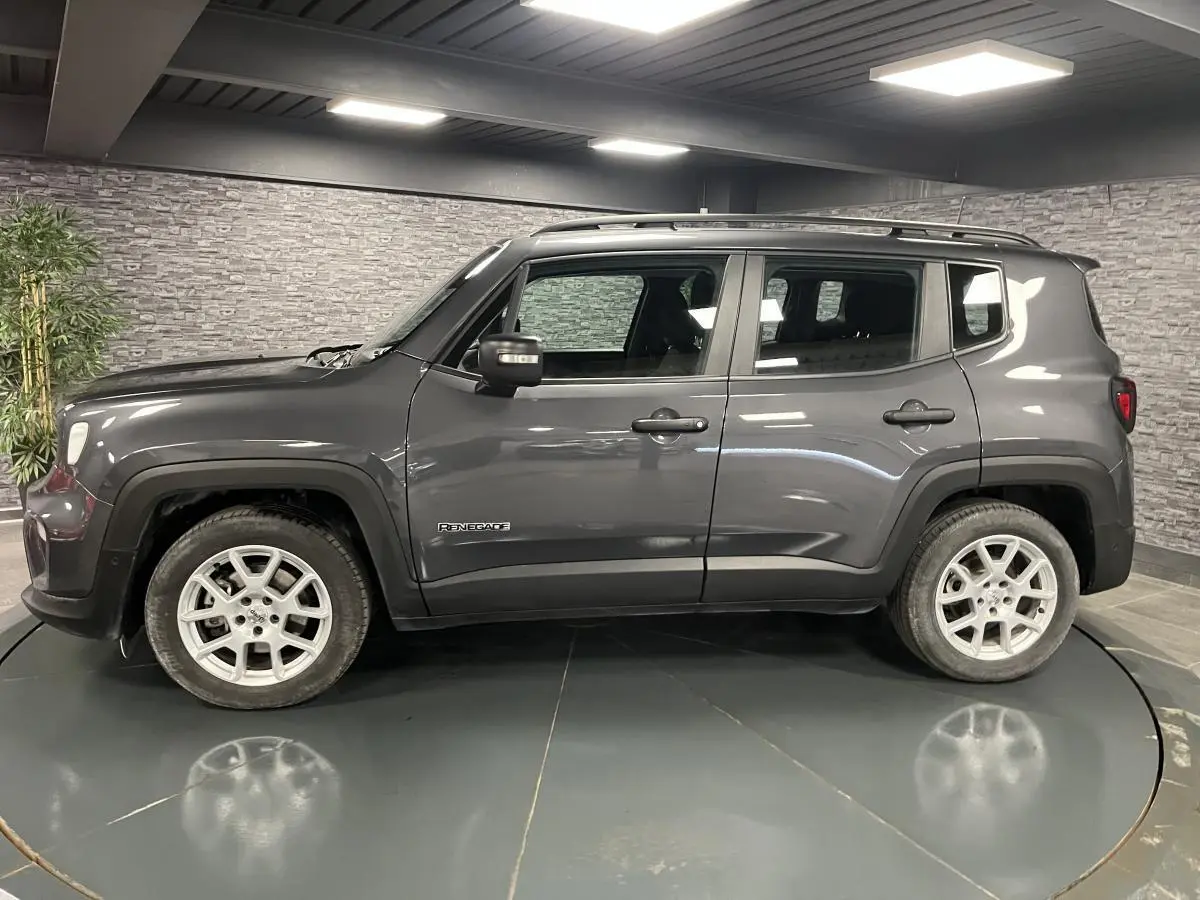 Vue de profil côté gauche d'un Jeep Renegade gris métallisé avec barres de toit noires en intérieur showroom.