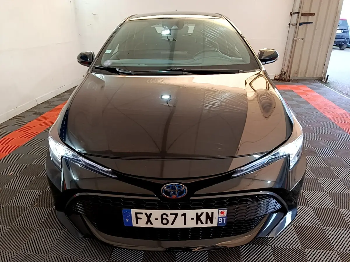 Vue avant d'une Toyota Corolla hybride noire 2021 avec calandre large et phares LED allumés dans un garage.