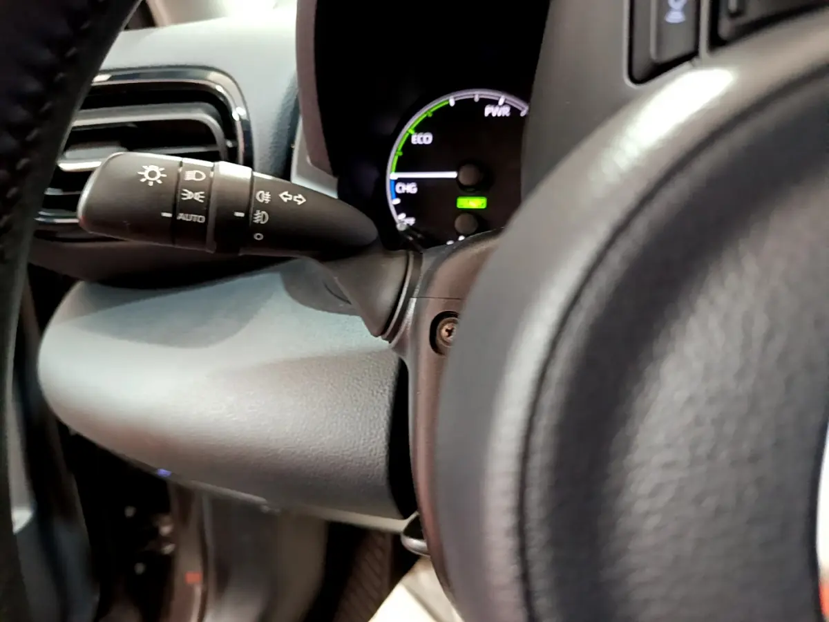 Gros plan sur le compteur hybride et le commodo d'éclairage à gauche du volant cuir d'une Toyota Yaris gris clair 2021.