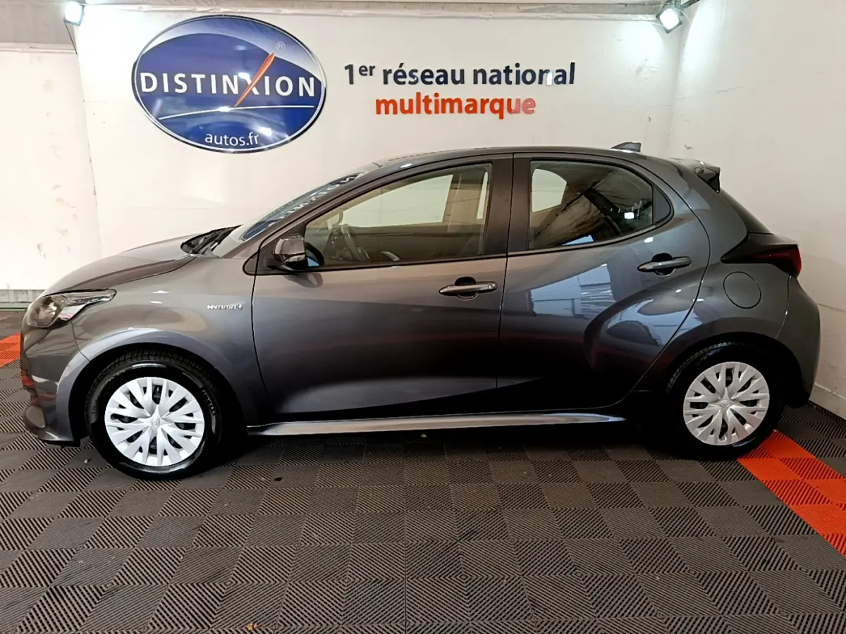Profil côté gauche d'une Toyota Yaris hybride gris clair 2021, avec jantes à enjoliveurs blancs et logo hybride visible.