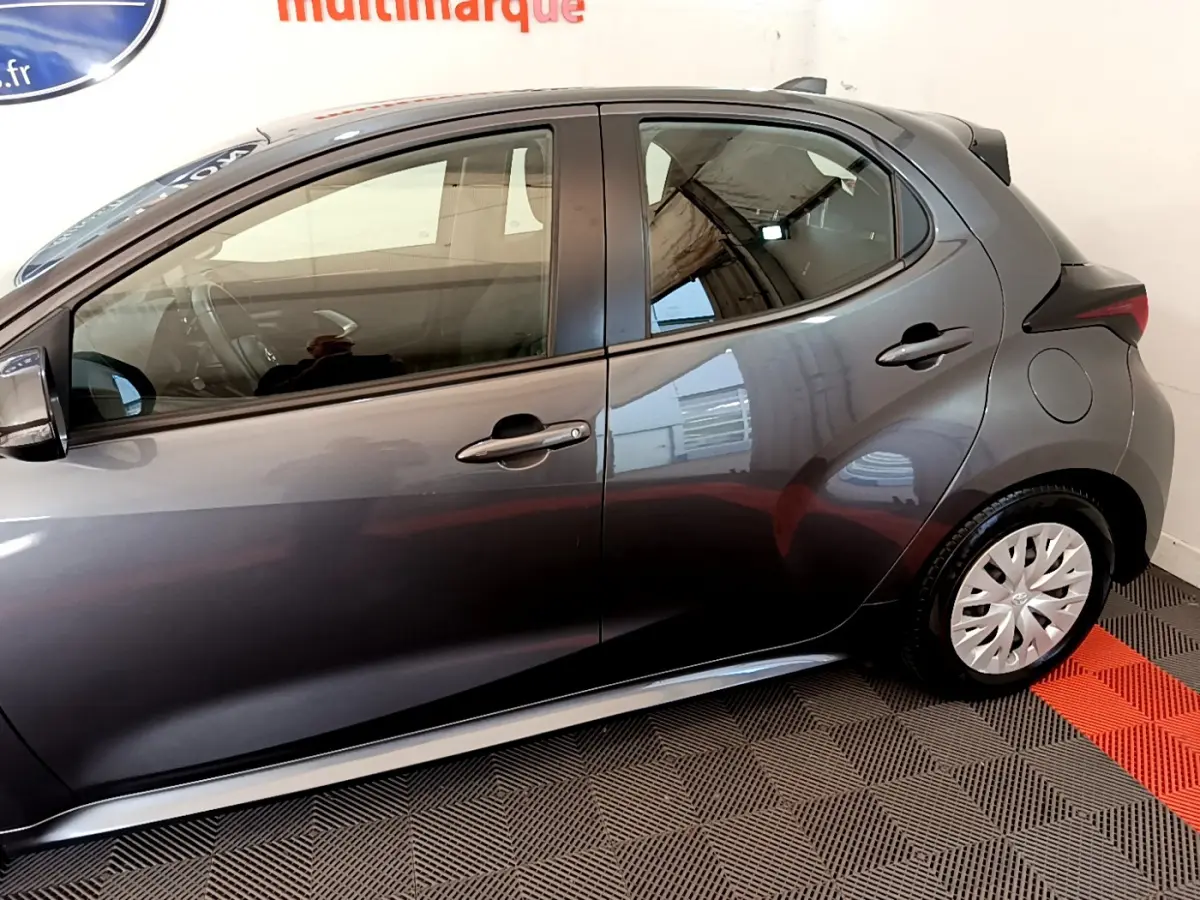 Profil côté gauche d'une Toyota Yaris hybride gris clair, avec jantes à enjoliveurs et poignées de porte assorties.
