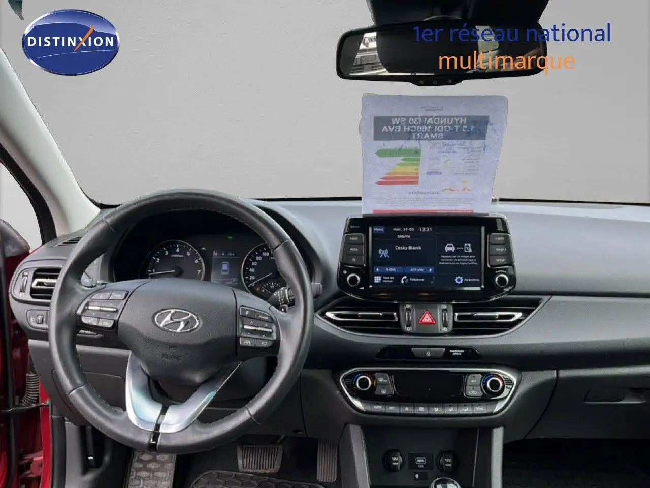 Vue intérieure frontale du tableau de bord et volant d’une Hyundai i30 SW 2023 en rouge Sunset Metal, avec écran tactile allumé.