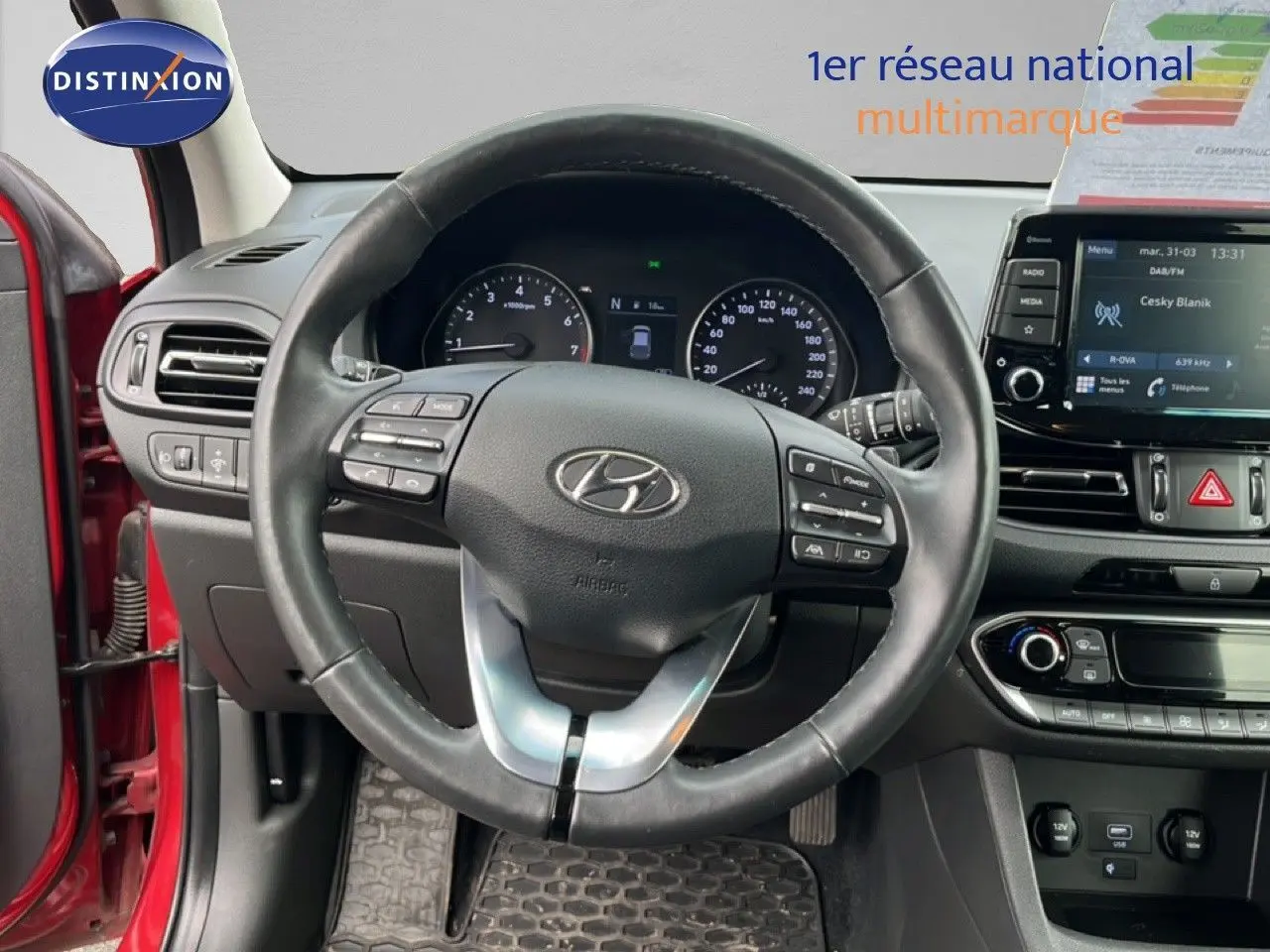 Vue intérieure centrée sur le volant cuir d’une Hyundai i30 SW rouge, avec tableau de bord et écran multimédia visibles.