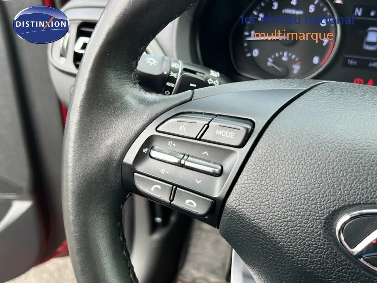 Gros plan sur le volant côté gauche de la Hyundai i30 SW 2023 en rouge Sunset Red, avec commandes audio intégrées.