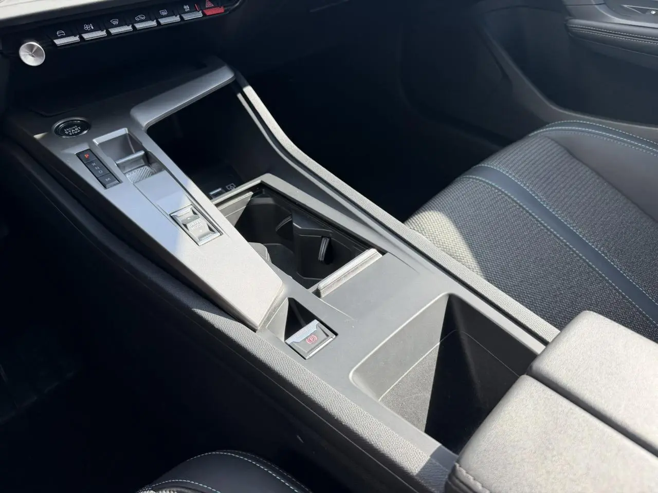 Vue intérieure rapprochée de la console centrale et accoudoir avant de la Peugeot 308 SW 2025, sièges tissu gris avec surpiqûres bleues.