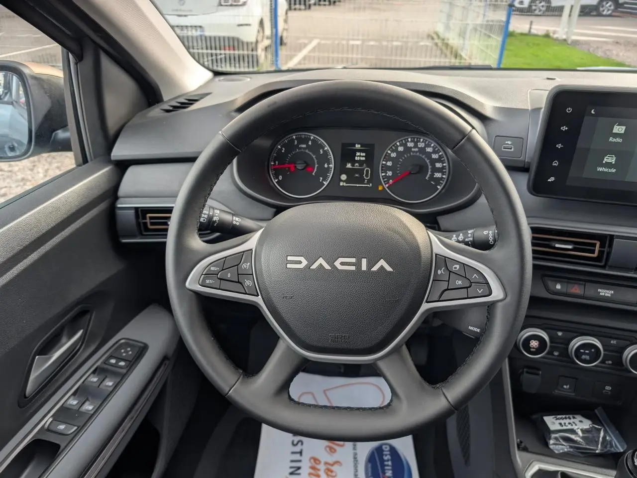 Vue intérieure centrée sur le volant DACIA noir avec tableau de bord et écran tactile à droite, portière côté gauche visible.