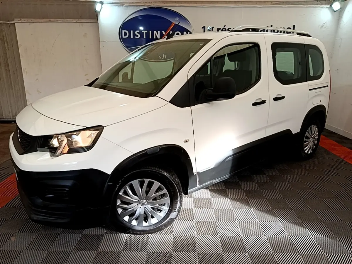 Vue 3/4 avant gauche d’un Peugeot Rifter blanc avec protections noires et jantes alliage, en intérieur showroom.