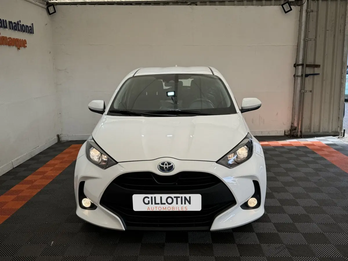 Vue frontale d'une Toyota Yaris hybride blanche 2022 avec phares allumés dans un garage showroom.