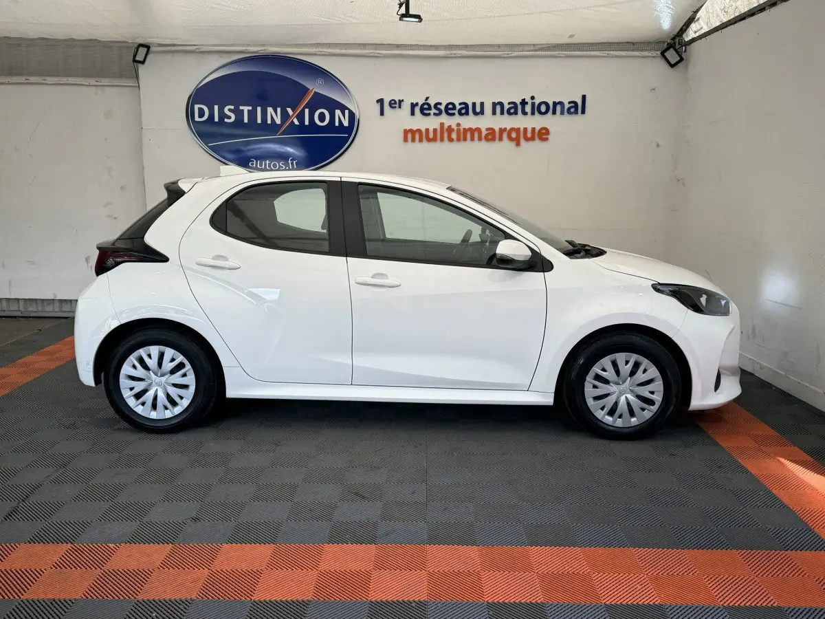 Profil côté gauche d'une Toyota Yaris hybride blanche 2022, avec jantes standard et toit noir contrasté en arrière-plan intérieur.