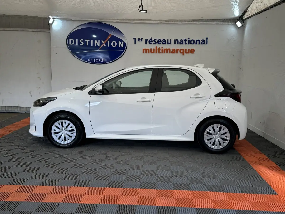 Profil côté gauche d'une Toyota Yaris blanche hybride 2022, avec jantes et feux arrière distinctifs visibles en intérieur showroom.