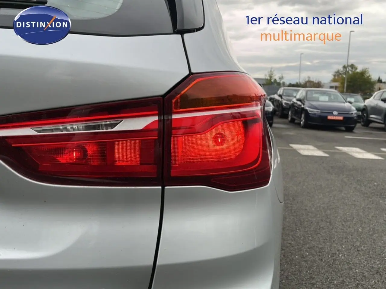 Gros plan sur le feu arrière droit d’un BMW X1 gris argent métal, avec un parking et plusieurs voitures en arrière-plan.