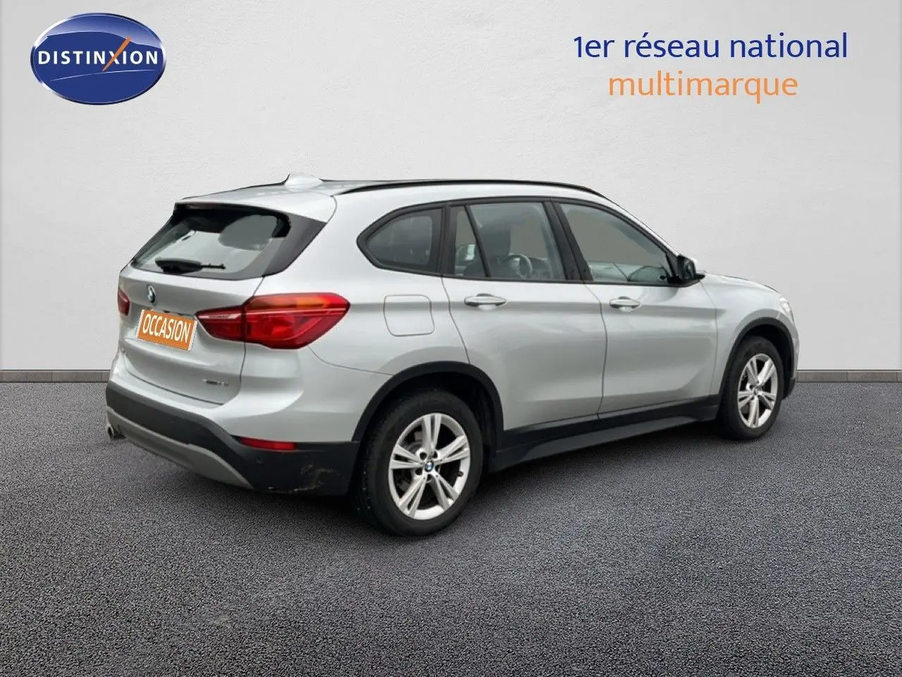 BMW X1 18i gris argent métal vue 3/4 arrière côté droit avec jantes alliage et feux arrière allumés.