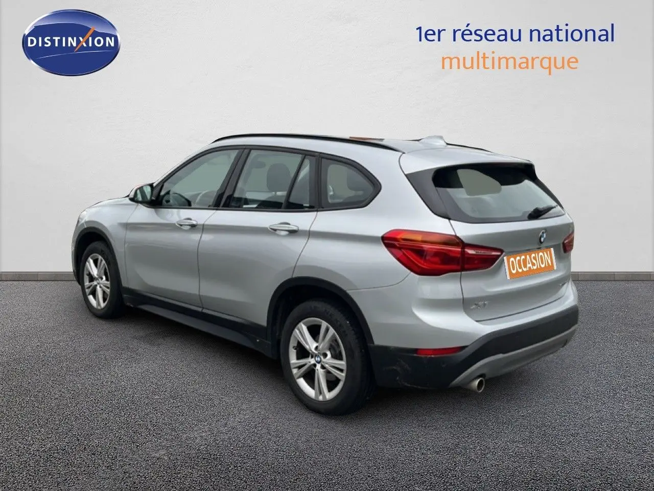 BMW X1 18i gris argent métal en 3/4 arrière droit, avec jantes alliage et feux arrière allumés.