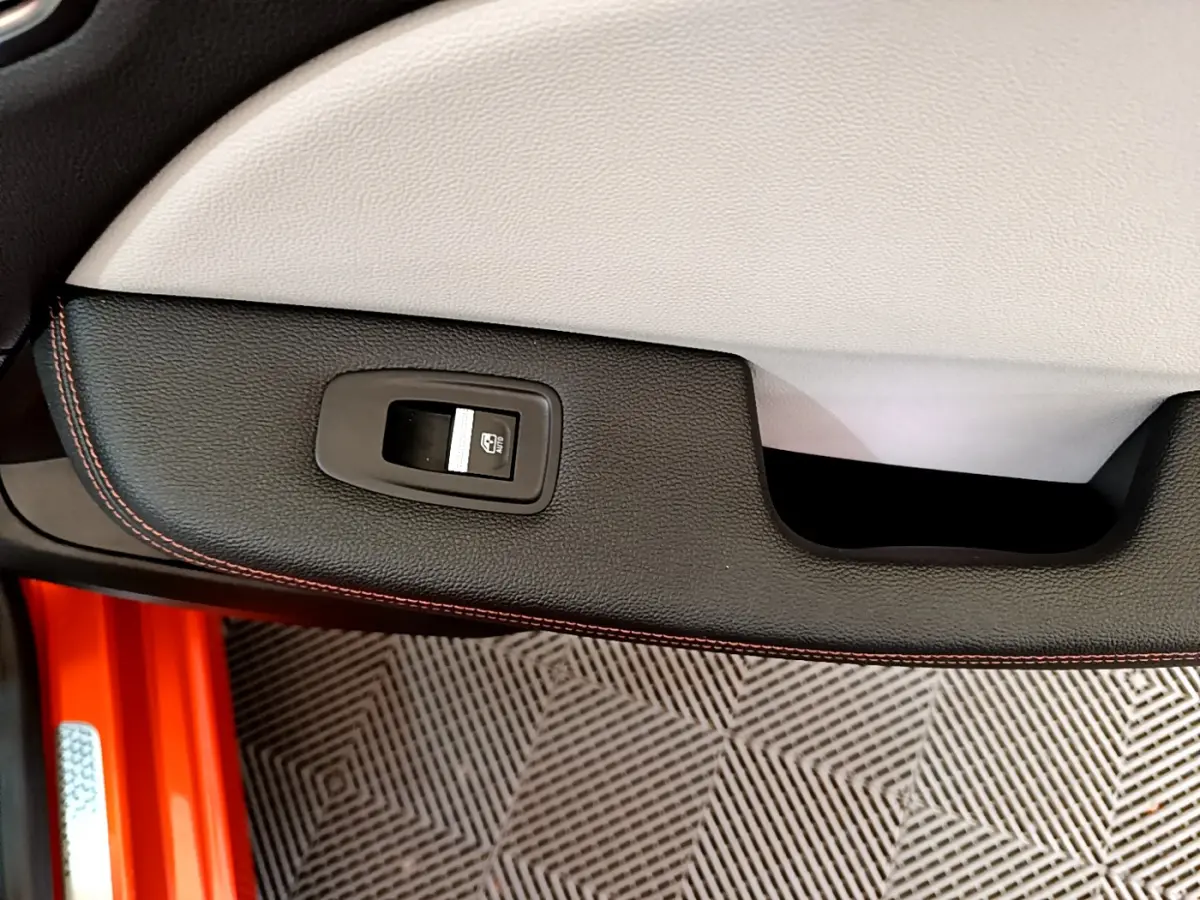 Gros plan sur la commande de lève-vitre côté gauche de la porte intérieure noire avec surpiqûres orange d'une Renault Clio orange.