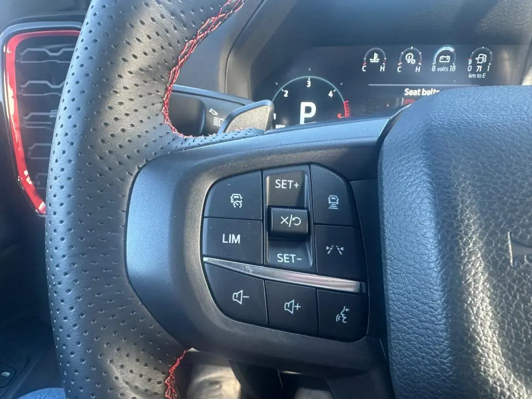 Gros plan sur les commandes à gauche du volant perforé noir avec surpiqûres rouges du Ford Ranger blanc, tableau de bord numérique en arrière-plan.