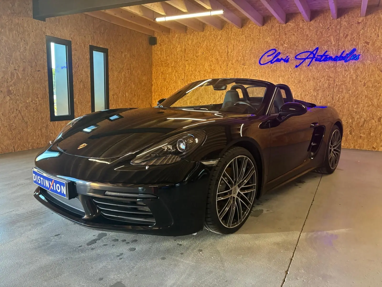 Porsche 718 Boxster 2.0 300 noir vue 3/4 avant droit en intérieur avec jantes alliage et capote ouverte.