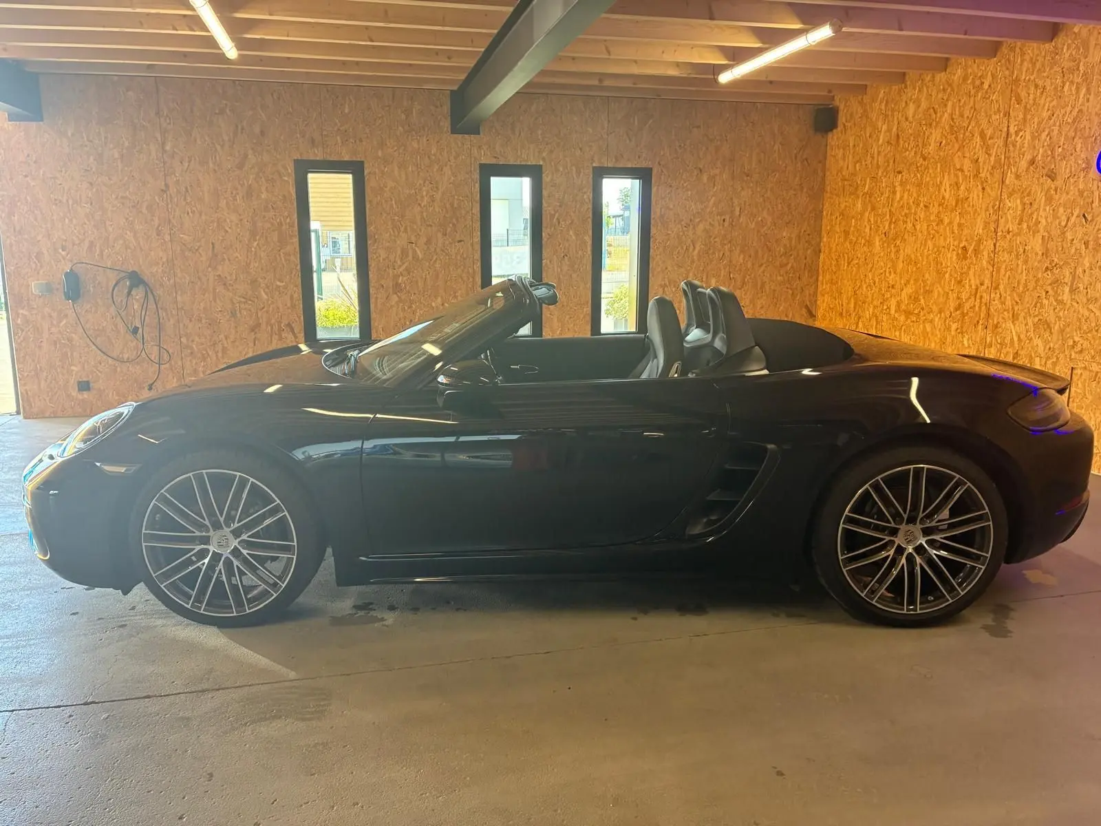 Vue de profil côté gauche d'une Porsche 718 Boxster noire cabriolet, avec jantes alliage et intérieur apparent.