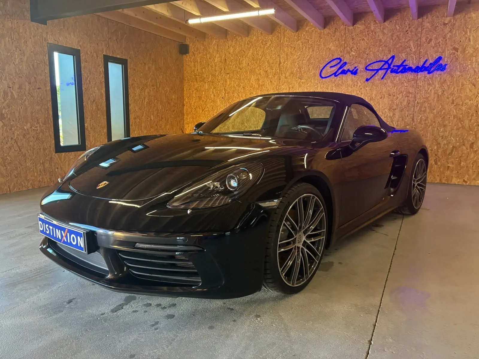 Porsche 718 Boxster noir vue 3/4 avant droit en intérieur, avec jantes alliage et capote fermée.