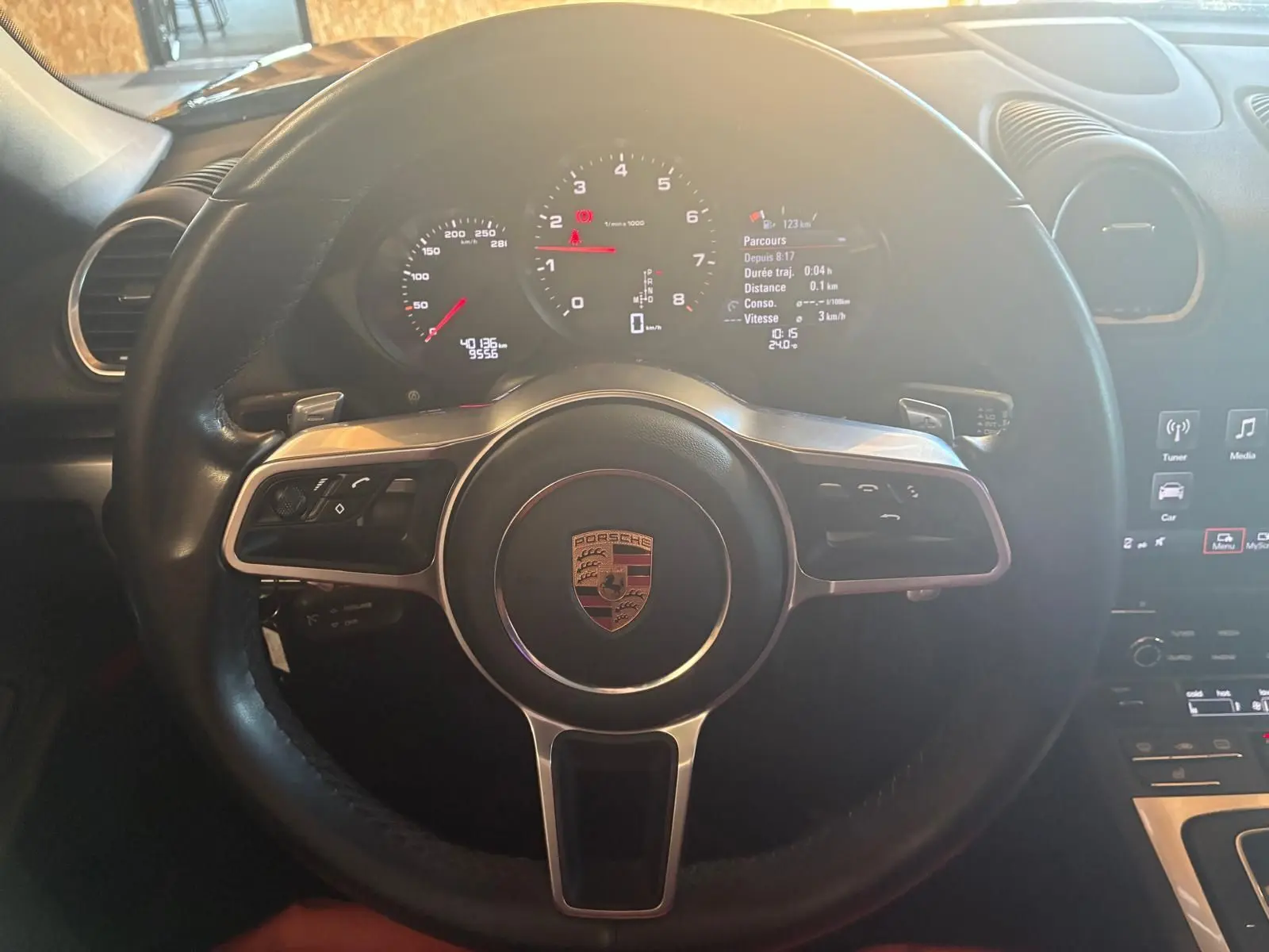 Vue rapprochée du volant noir de la Porsche 718 Boxster 2.0 300 de 2018 avec tableau de bord numérique allumé.