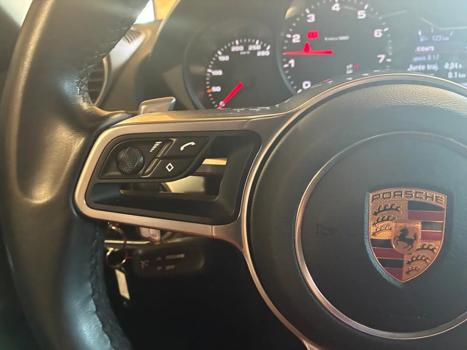 Gros plan sur le volant noir d’un Porsche 718 Boxster 2.0 300 de 2018 avec commandes intégrées et tableau de bord lumineux.