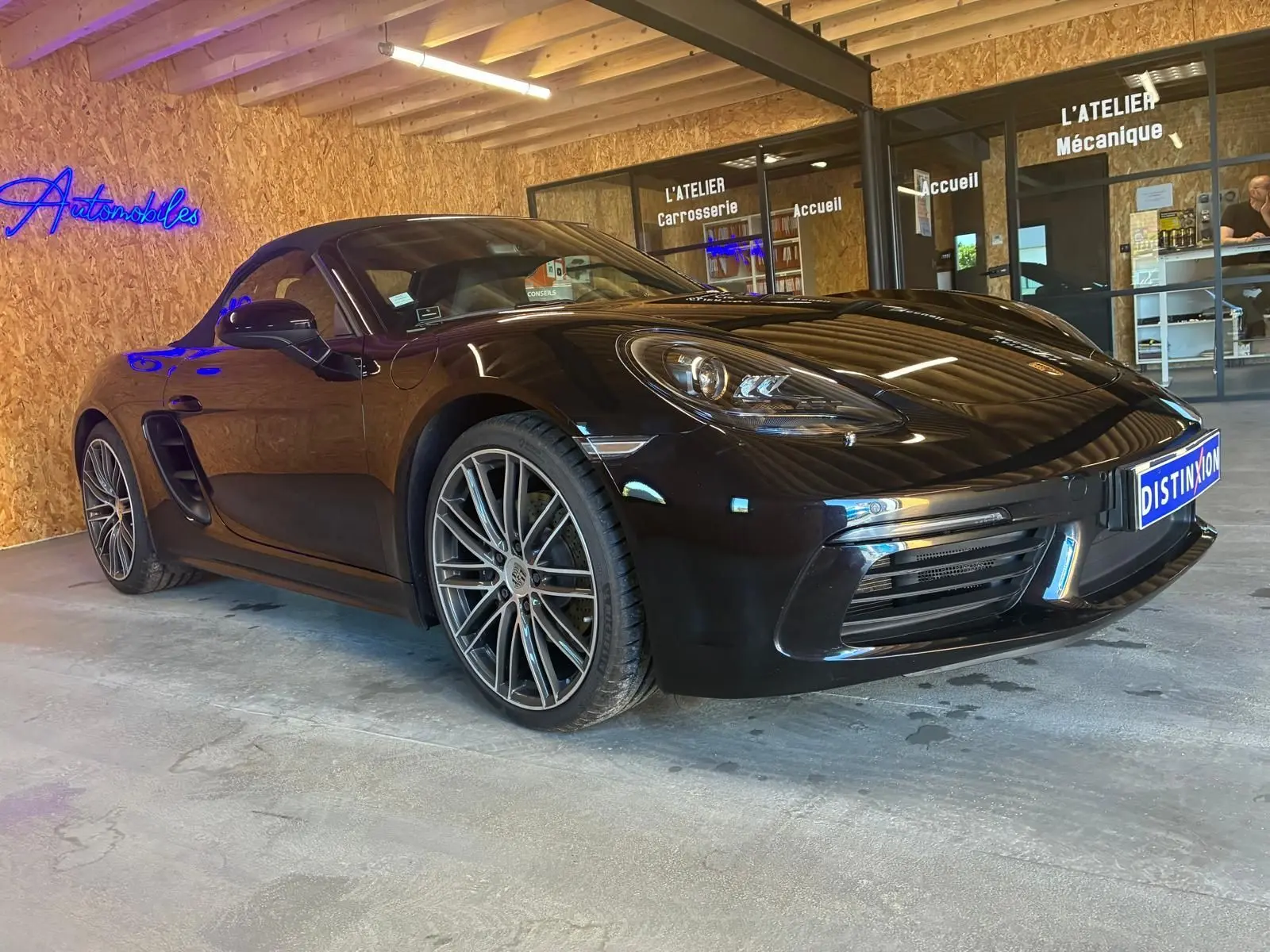 Porsche 718 Boxster noir vue 3/4 avant droit, avec jantes multibranches et phares LED dans un garage.