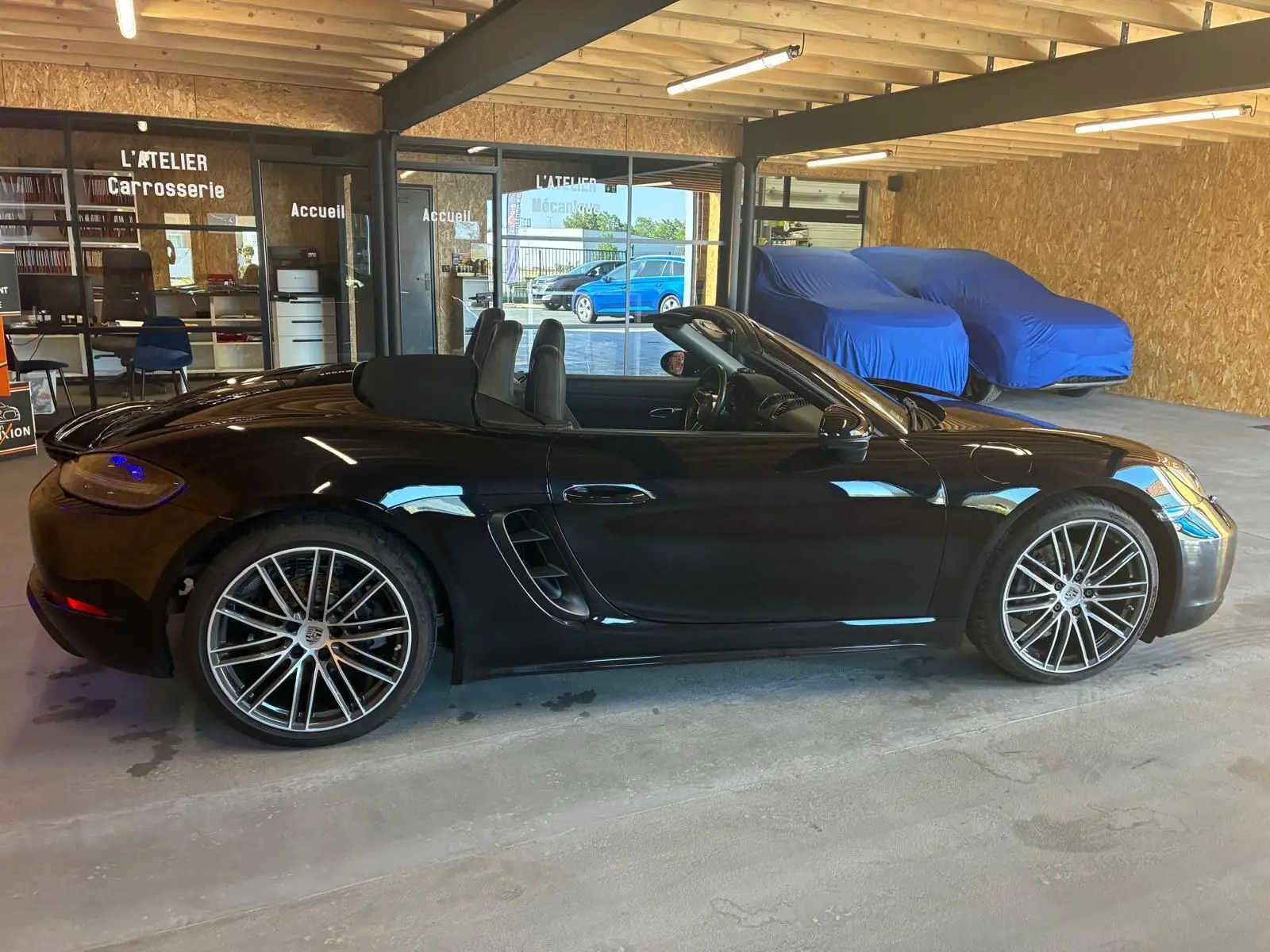 Vue de profil côté gauche d'une Porsche 718 Boxster noire cabriolet, avec jantes alliage et intérieur visible.