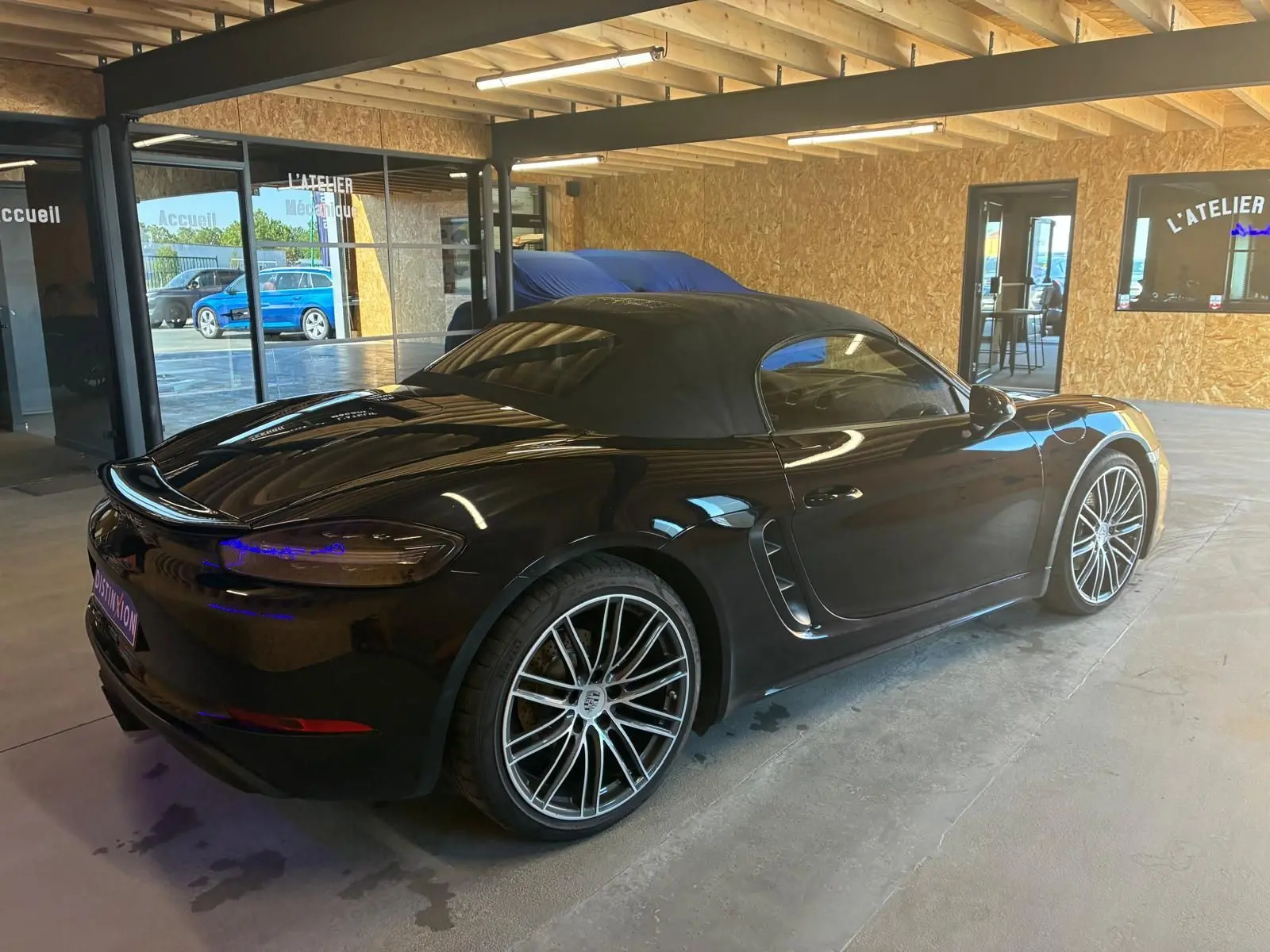 Porsche 718 Boxster 2.0 300 noir vue 3/4 arrière droit en intérieur avec toit souple fermé et jantes multibranches.