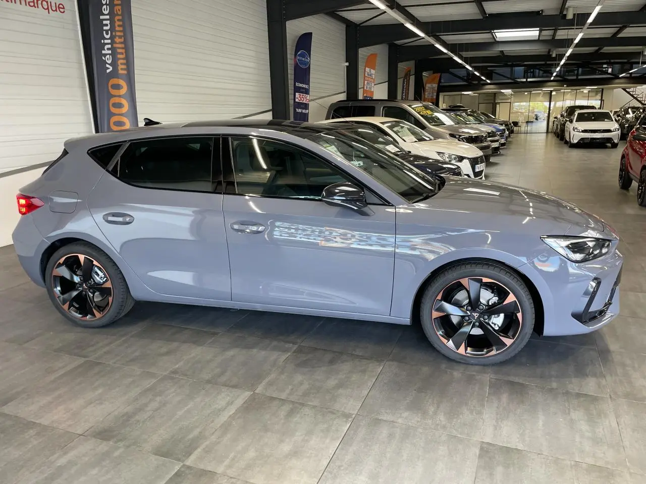 Vue latérale droite d'une CUPRA Leon 2026 gris Graphène avec jantes alliage noires et cuivrées en showroom.