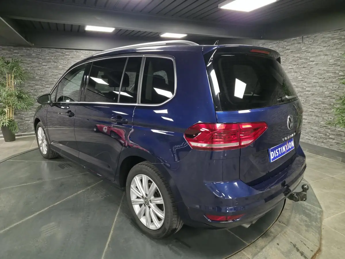 Vue 3/4 arrière droite d'un Volkswagen Touran bleu avec attelage escamotable en intérieur showroom.