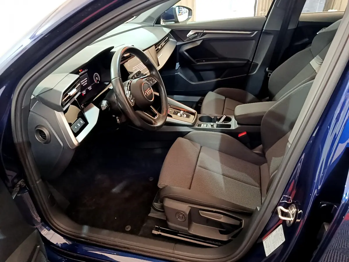 Intérieur côté conducteur d'une Audi A3 bleu 2023, volant cuir, tableau de bord digital et sièges tissu gris.