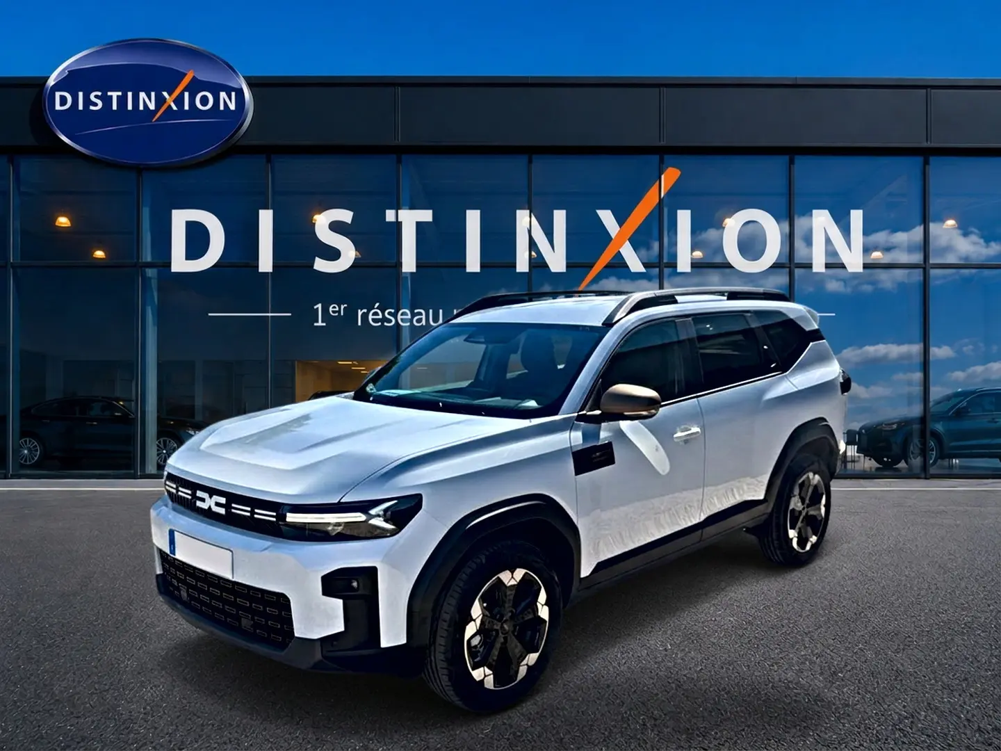 Dacia Bigster blanc en 3/4 avant droit, avec barres de toit et jantes alu distinctives, devant showroom Distinxion.
