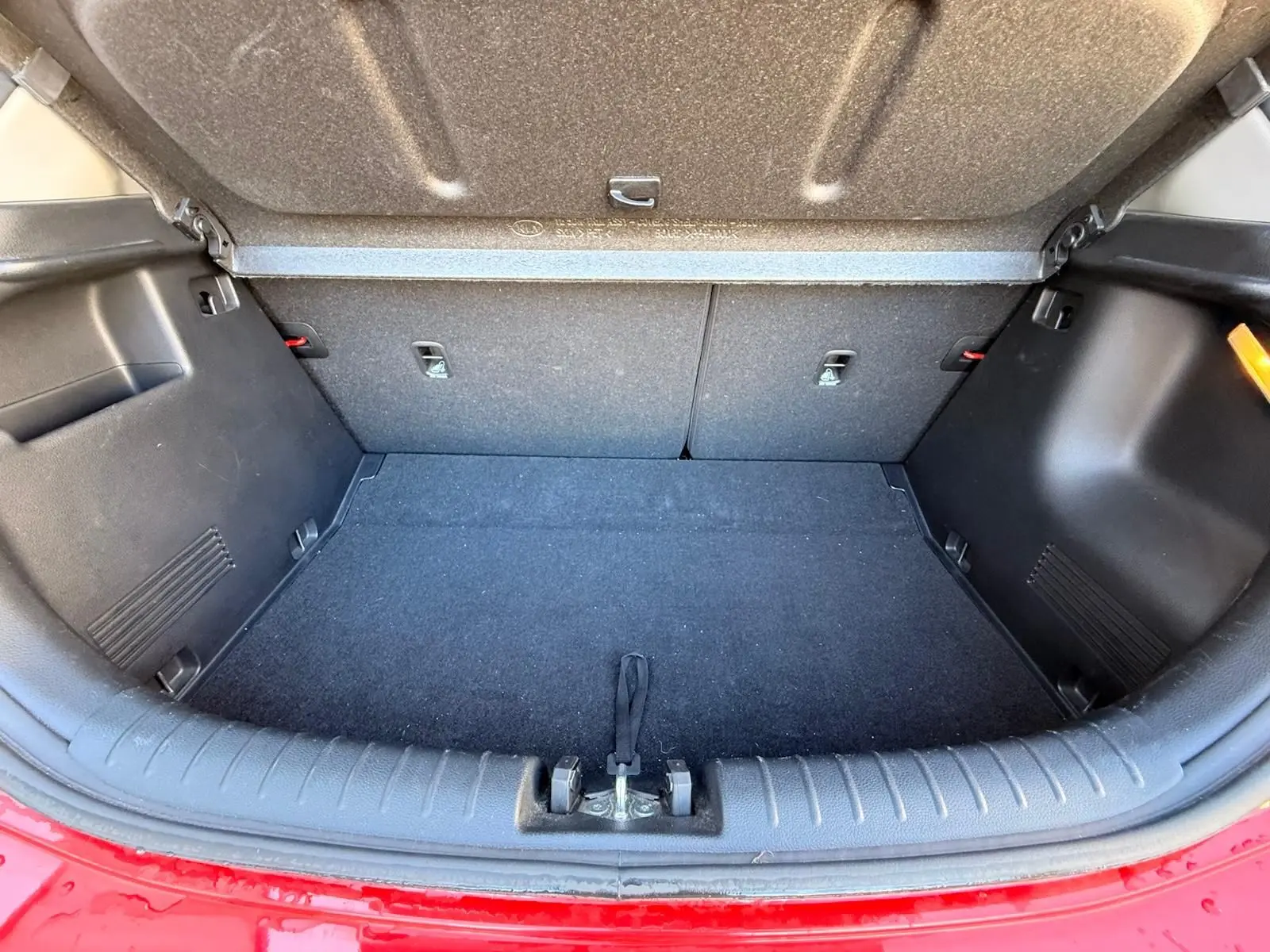 Coffre ouvert vu de l'arrière d'une KIA RIO rouge grenadine, montrant l'espace de chargement vide et les finitions intérieures noires.