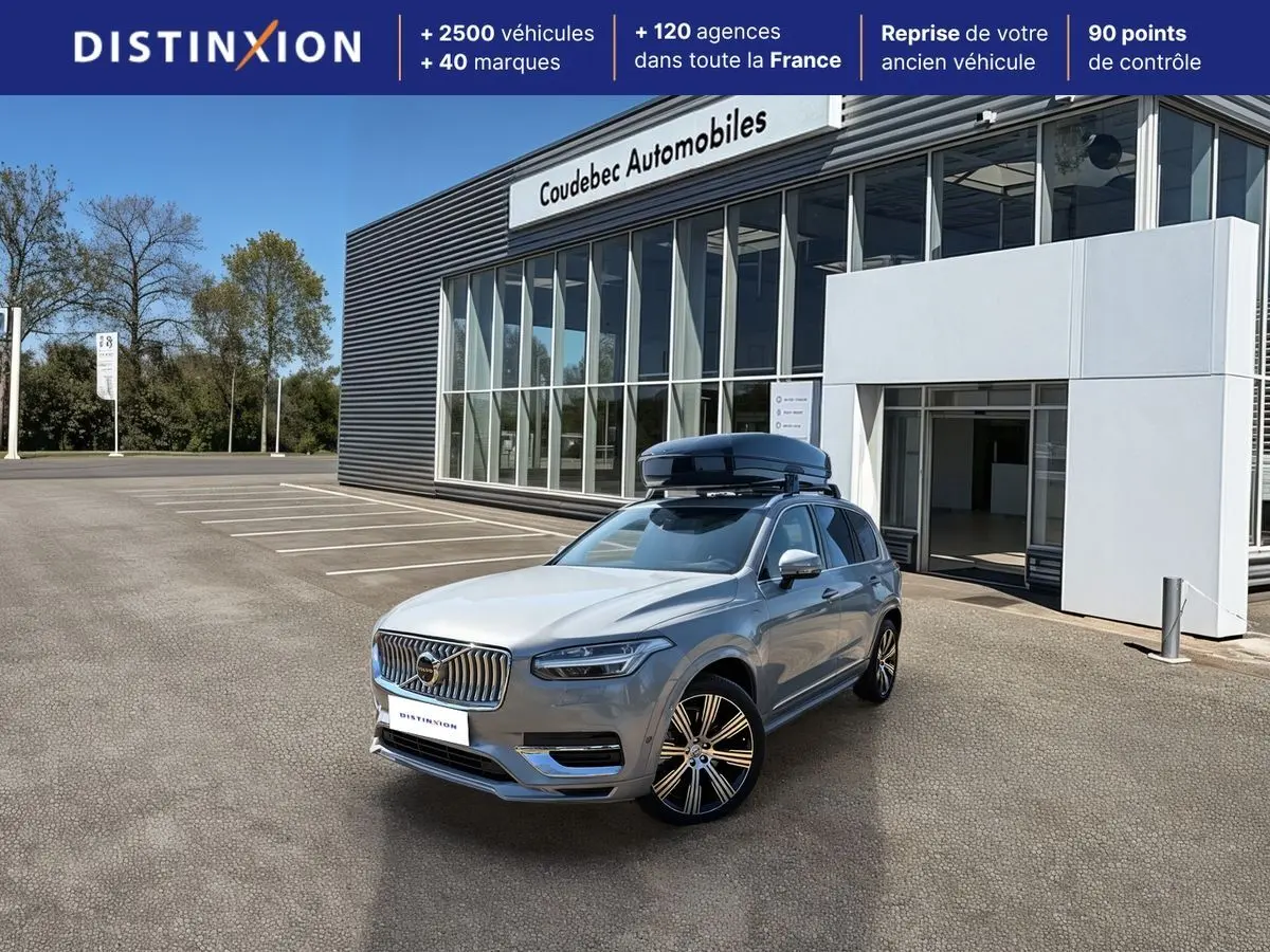 Vue 3/4 avant droite du Volvo XC90 gris avec coffre de toit noir sur parking devant concession.