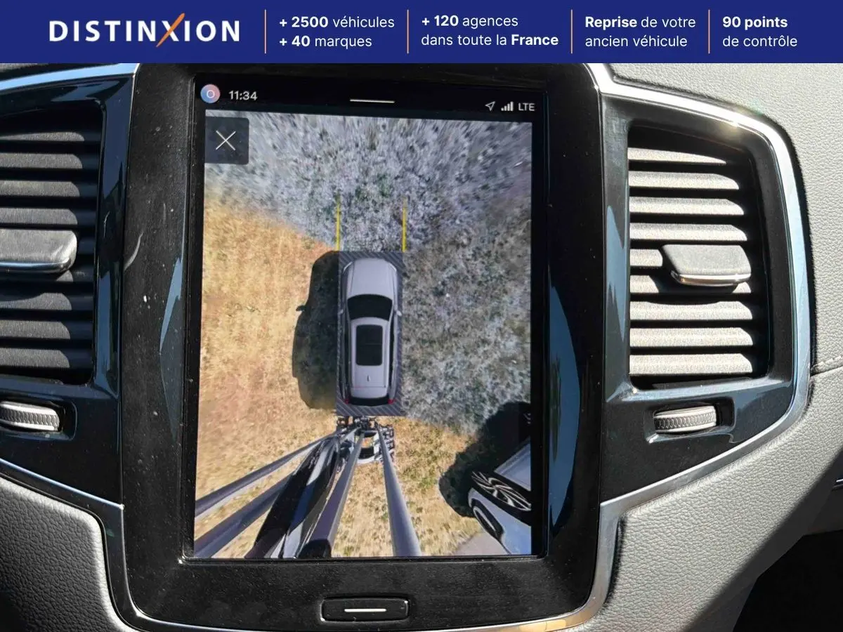 Vue intérieure de l'écran central montrant la caméra surround view 360° d'un Volvo XC90 gris stationné sur un sol mixte.