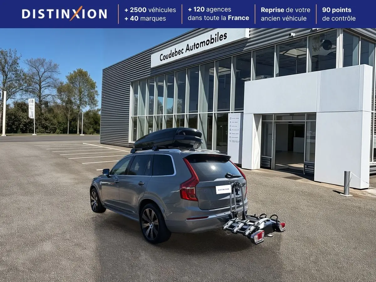Vue 3/4 arrière d'un Volvo XC90 gris avec coffre de toit noir et porte-vélos fixé à l'attelage, devant un bâtiment commercial.