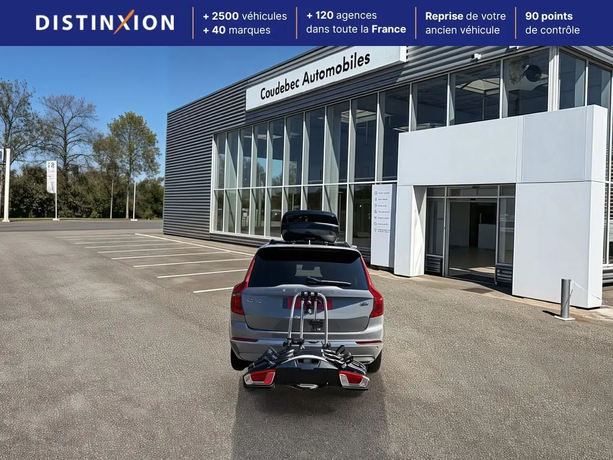 Vue arrière d'un Volvo XC90 gris avec coffre de toit et porte-vélos, stationné devant un bâtiment commercial.