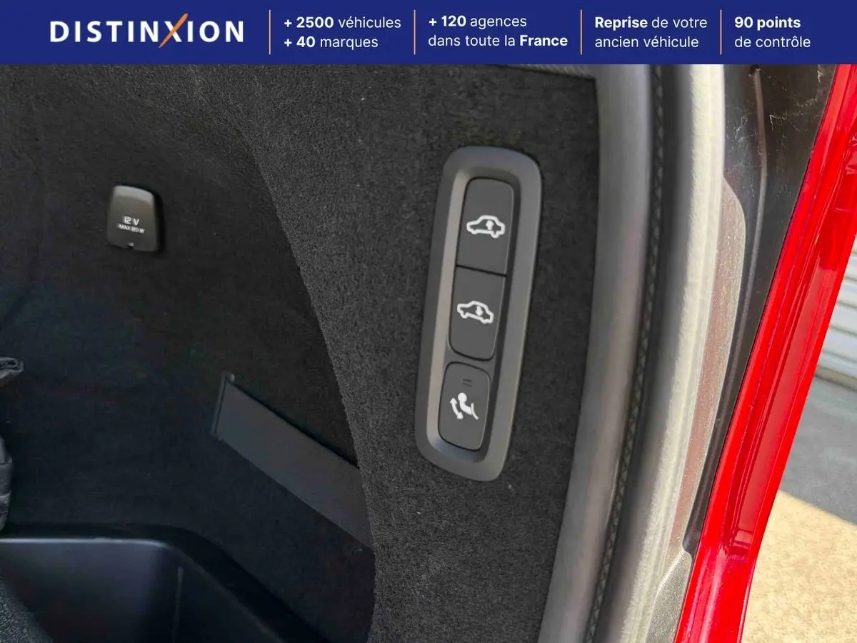 Gros plan sur les commandes intérieures du coffre d'un Volvo XC90 gris, avec bouton rabattage sièges et prise 12V visible.