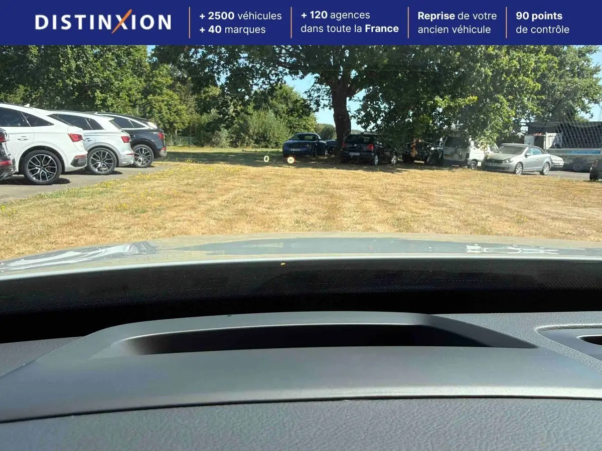 Vue depuis l'intérieur du Volvo XC90 gris 2024, montrant le tableau de bord et le pare-brise sur un parking extérieur.