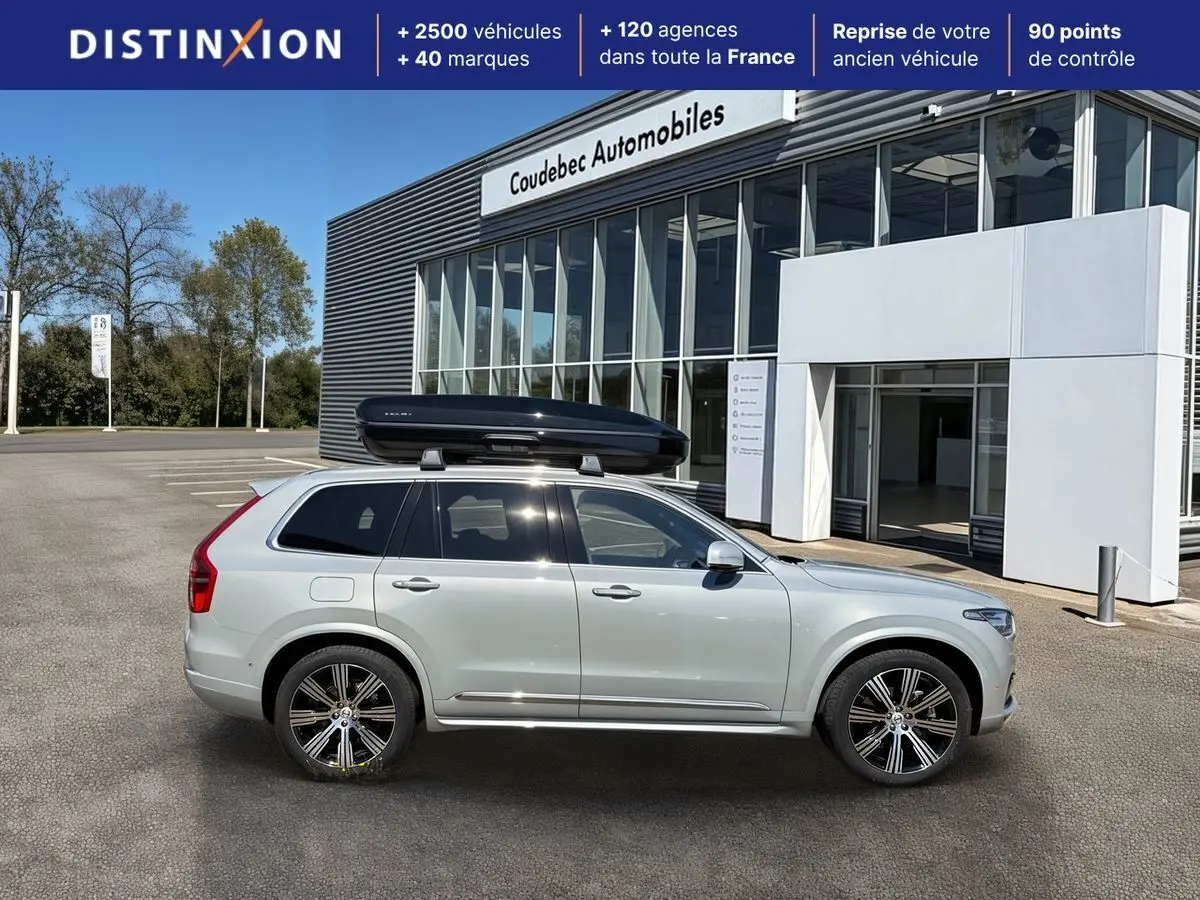 Vue de profil droit du Volvo XC90 gris clair avec coffre de toit noir sur un parking devant un bâtiment commercial.