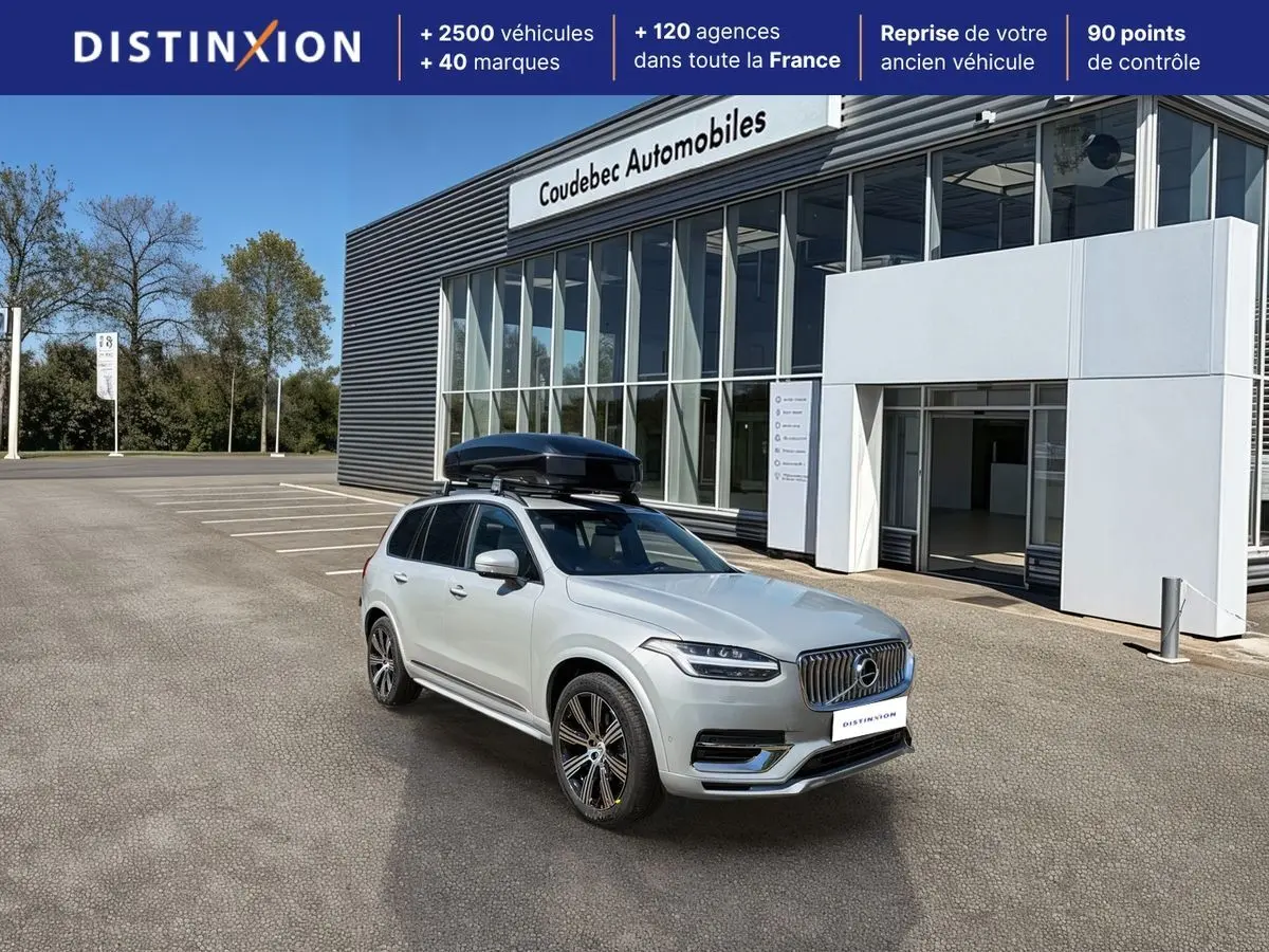 Vue 3/4 avant droite du Volvo XC90 gris avec coffre de toit noir, stationné devant un bâtiment commercial.