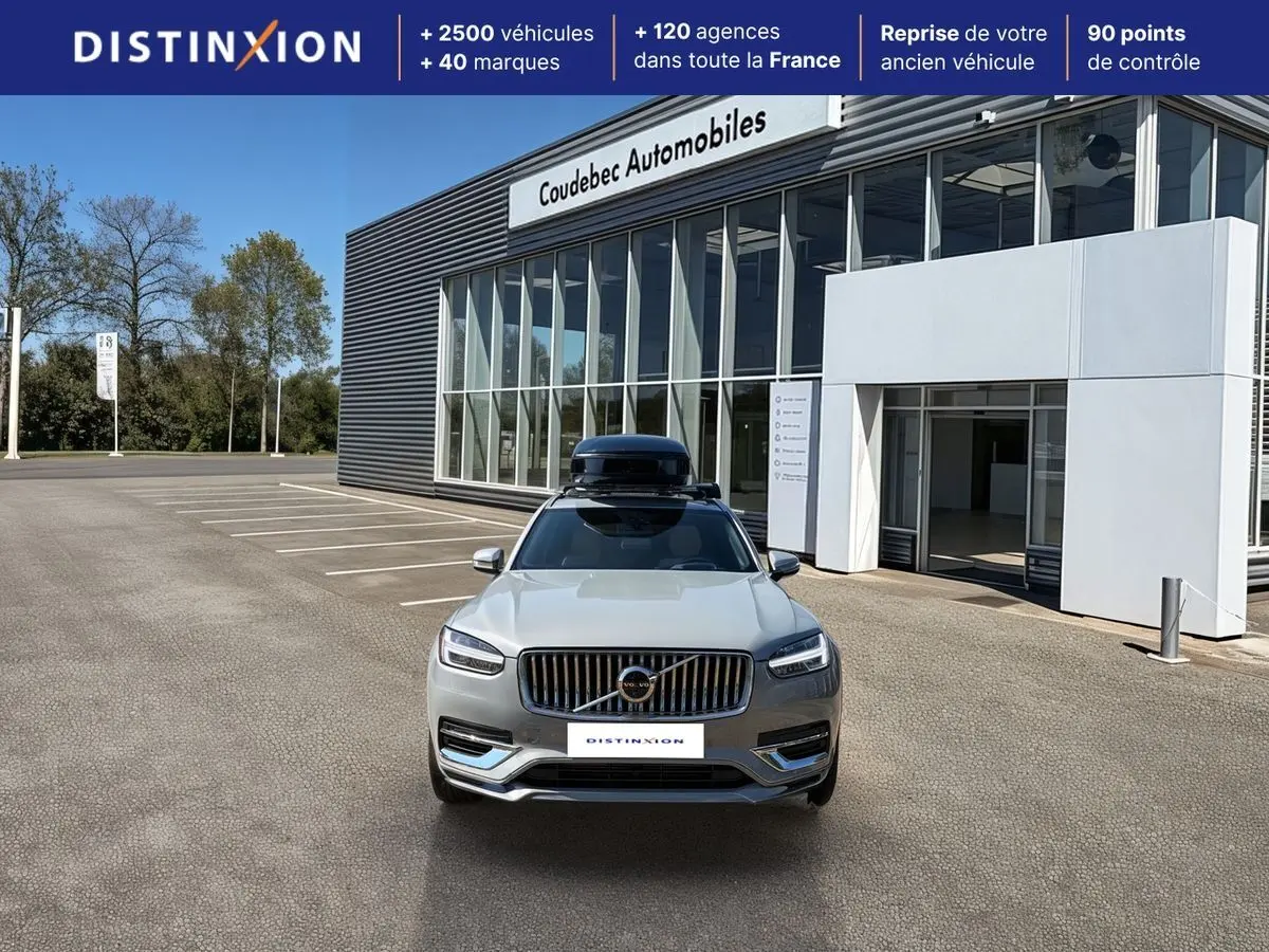Vue avant d'un Volvo XC90 gris avec coffre de toit noir, stationné devant un bâtiment commercial.