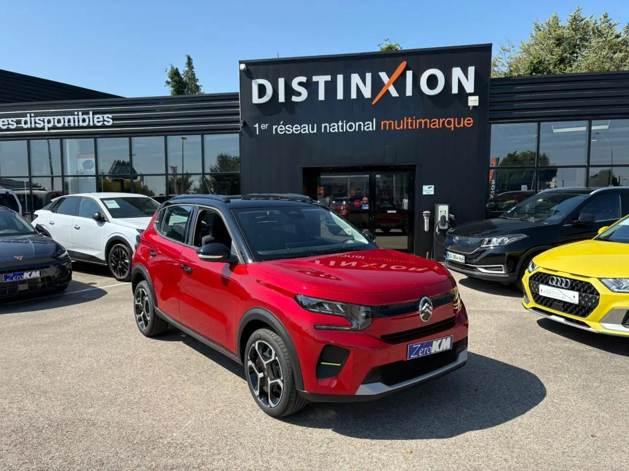Citroën C3 2025 rouge Elixir avec toit noir, vue 3/4 avant droit, stationnée devant un concessionnaire multimarque.