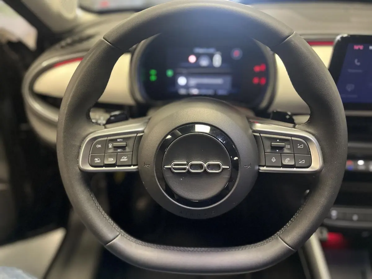 Gros plan sur le volant noir du Fiat 600 La Prima 2024 avec logo 600 et commandes intégrées, planche de bord ivoire en arrière-plan.