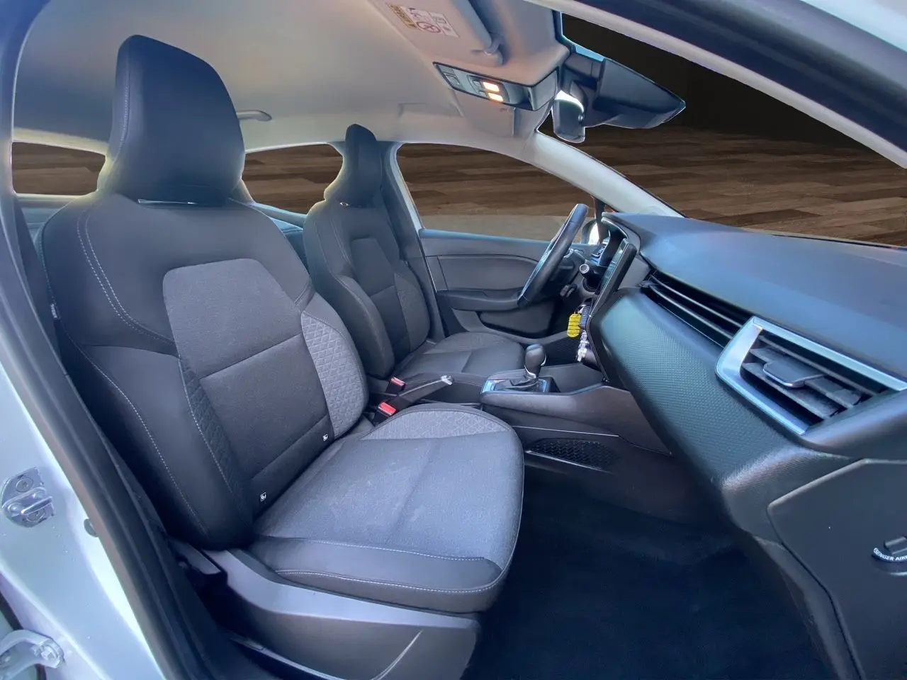 Vue intérieure côté gauche de la Renault Clio V blanche, montrant les sièges noirs et la planche de bord moderne.