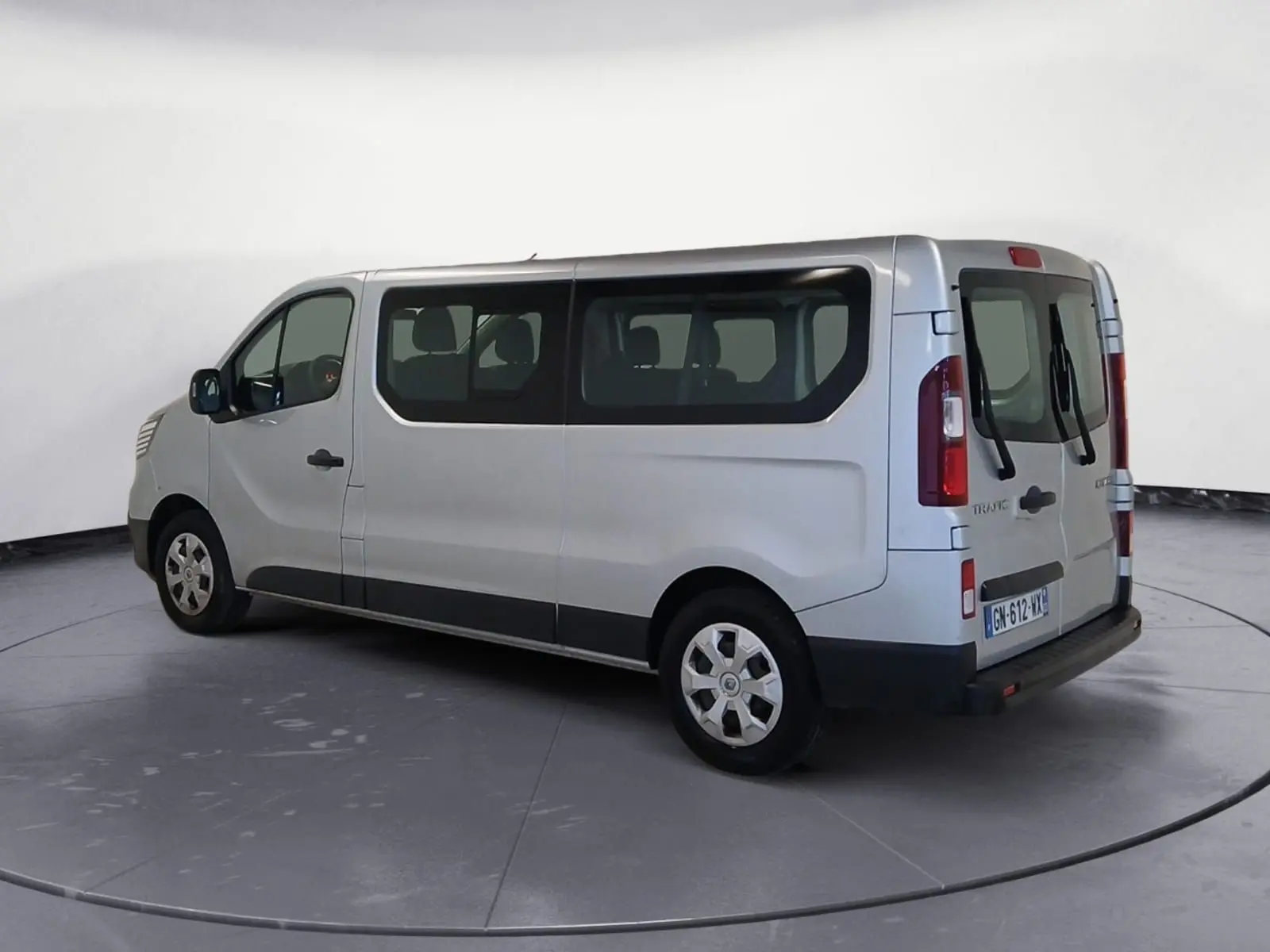Vue 3/4 arrière droite d’un Renault Trafic Combi gris clair métal, montrant ses grandes fenêtres latérales et ses portes arrière vitrées.