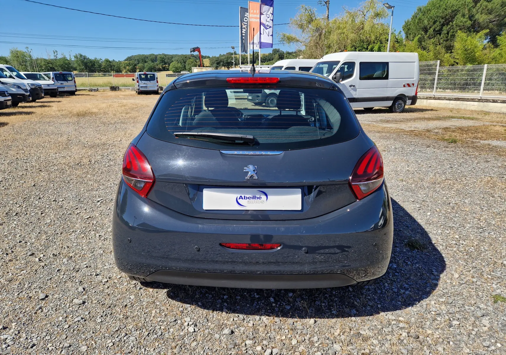 Vue arrière d'une Peugeot 208 gris hurricane 2018 garée sur un sol gravillonné avec plusieurs véhicules utilitaires en arrière-plan.