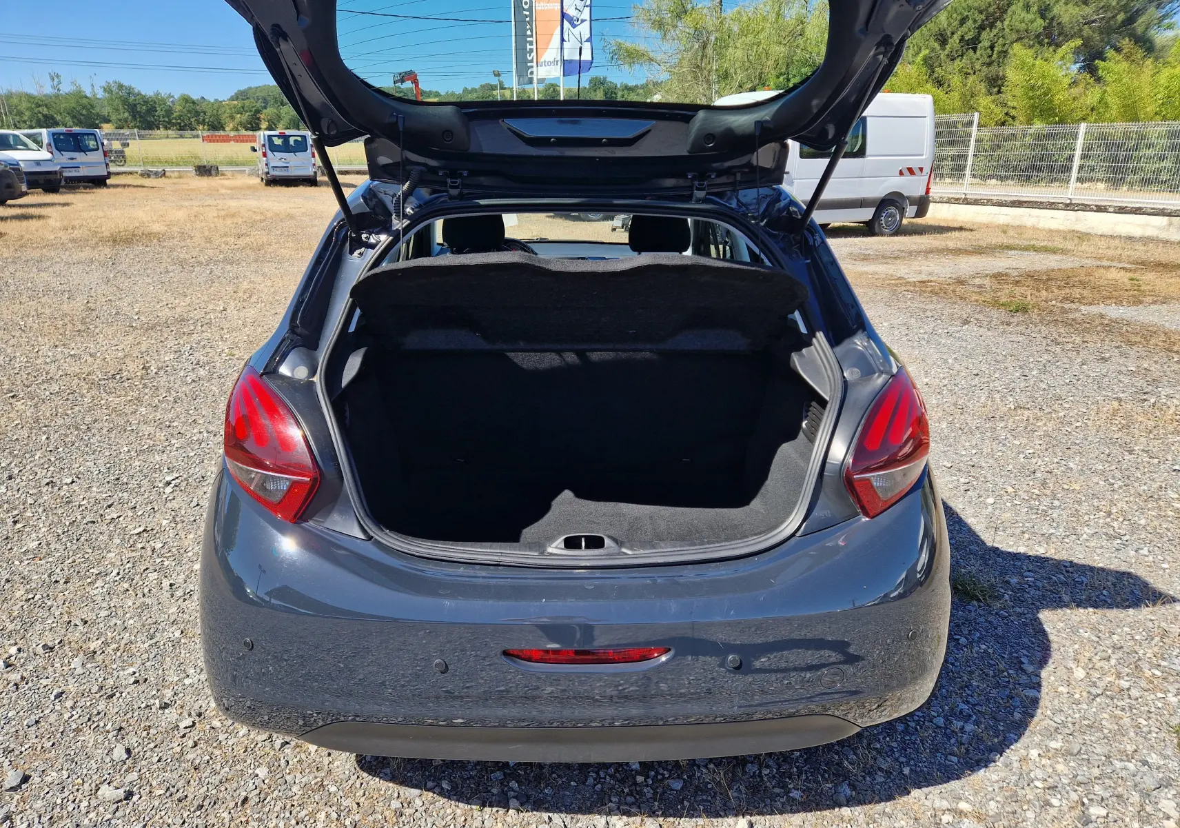 Vue arrière d'une Peugeot 208 gris hurricane de 2018 avec coffre ouvert sur un terrain extérieur ensoleillé.