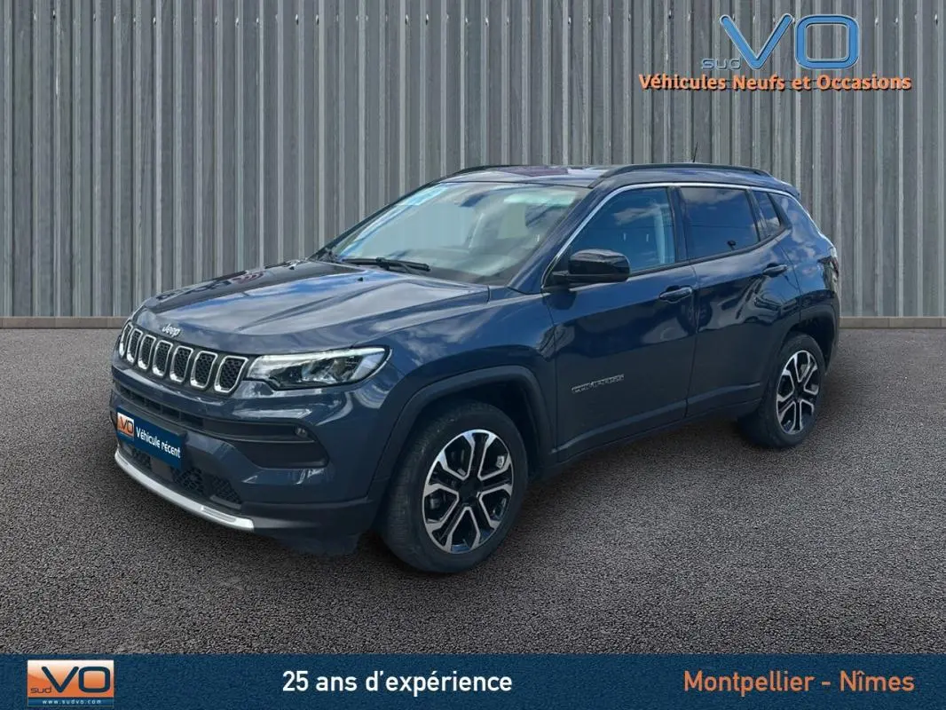 Jeep Compass bleu en 3/4 avant droit, avec barres de toit et jantes 18 pouces visibles sur sol bitumé.